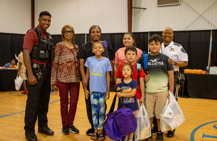 Group of children and adults posing with police officers at a community event, holding bags and backpacks.